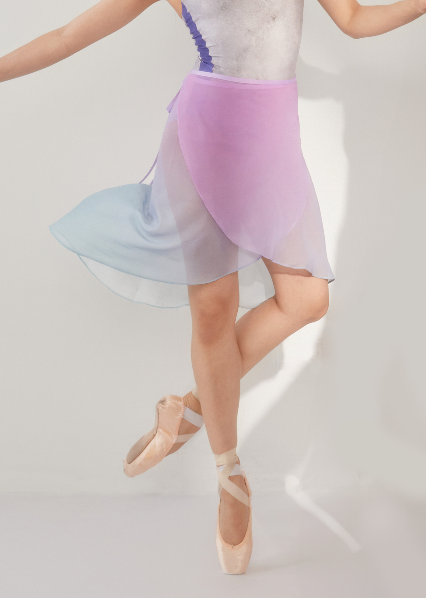 A ballet dancer in a lavender and blue ombre wrap skirt with a chiffon fabric, performing a dance move on pointe.