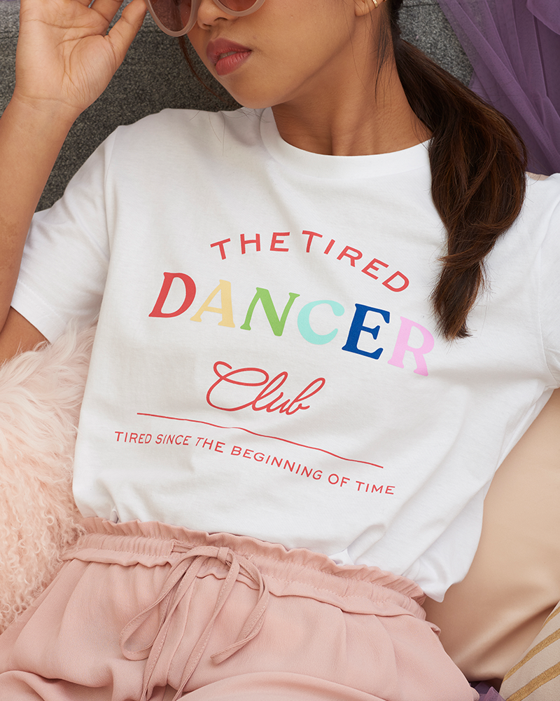 Person wearing a white t-shirt with colorful text 'THE TIRED DANCER Club' and pink pants.