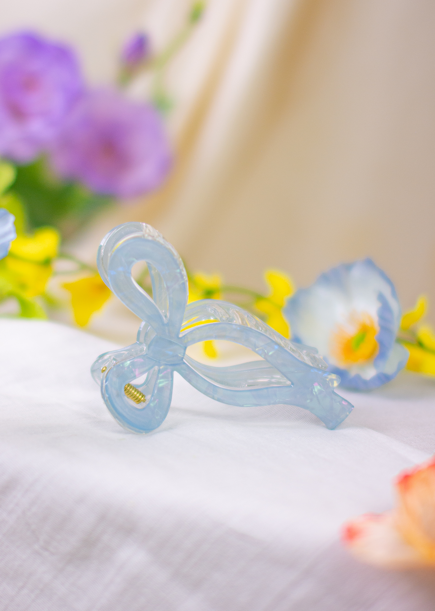 A blue ribbon-shaped acrylic claw clip placed on a white fabric surface with a blurred floral background.