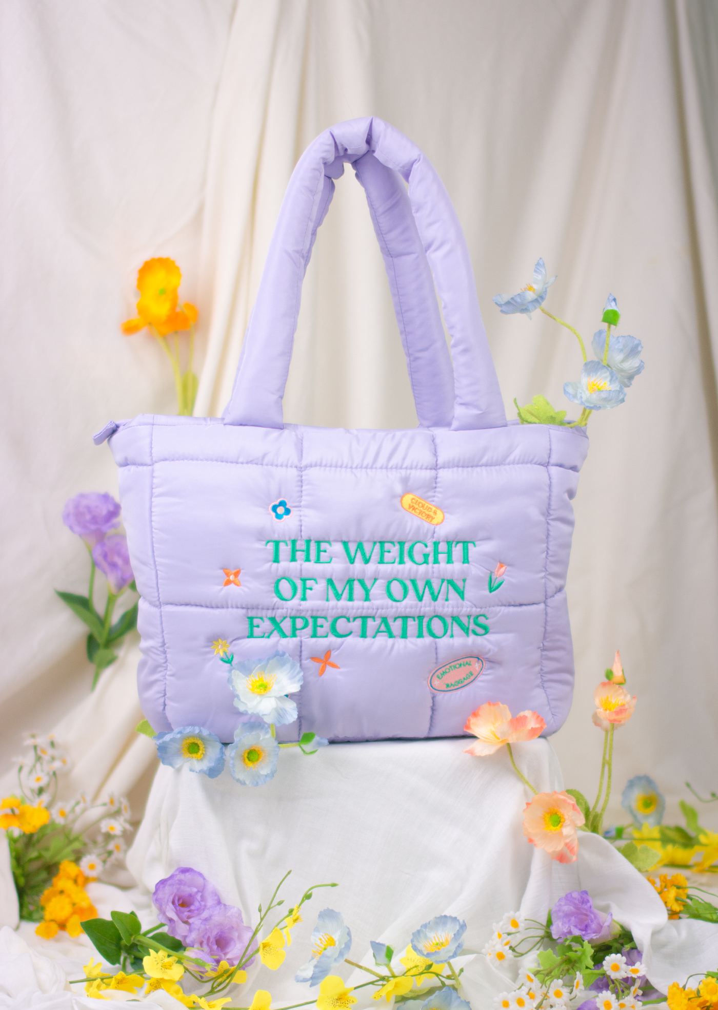 A lilac-colored puffer tote bag with embroidery on the front, displayed with flowers around it.